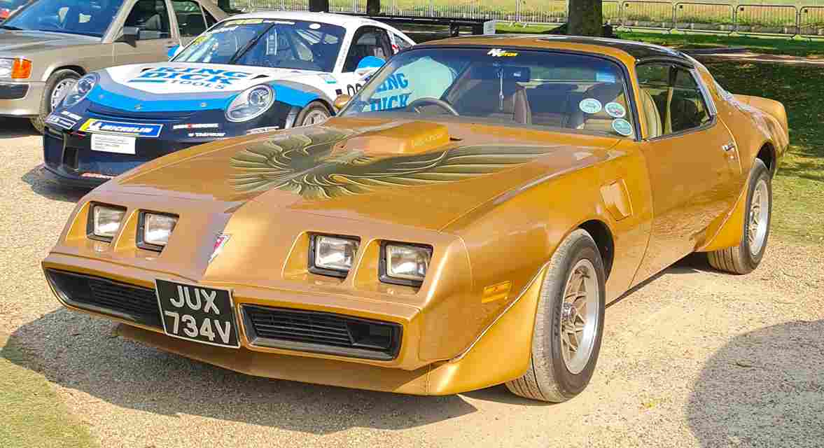 Golden-yellow vintage muscle car, likely a Pontiac Firebird Trans Am, with distinctive pop-up headlights and aggressive front design. Parked in what appears to be a display or show setting with other vehicles in background