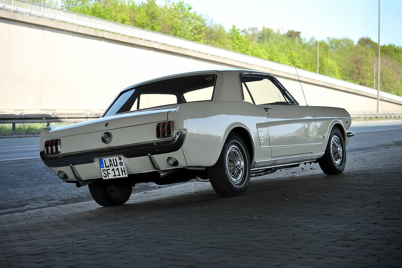 A vintage white 1966 Ford Mustang photographed from the rear three-quarter angle on a cobblestone surface. The classic sports car features distinctive triple taillights and a European license plate, with a concrete barrier and greenery in the background