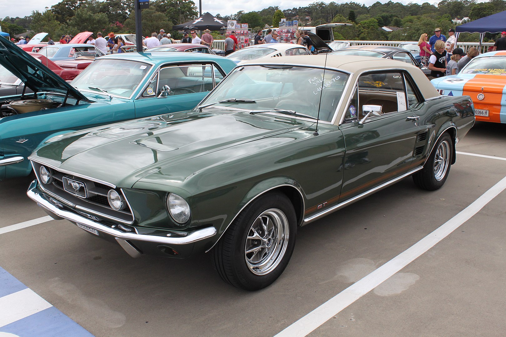 Dark green vintage Ford Mustang GT parked at a classic car show, with chrome details and polished wheels. Other classic cars and event attendees visible in the background on a sunny day