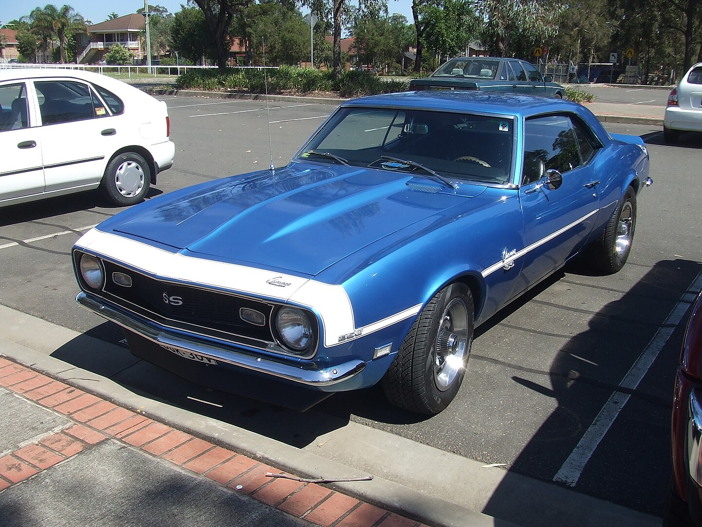 Classic blue Chevrolet Camaro SS with white racing stripes, parked in an urban parking lot. Vintage muscle car with gleaming paint, chrome details, and performance-oriented design surrounded by modern vehicles