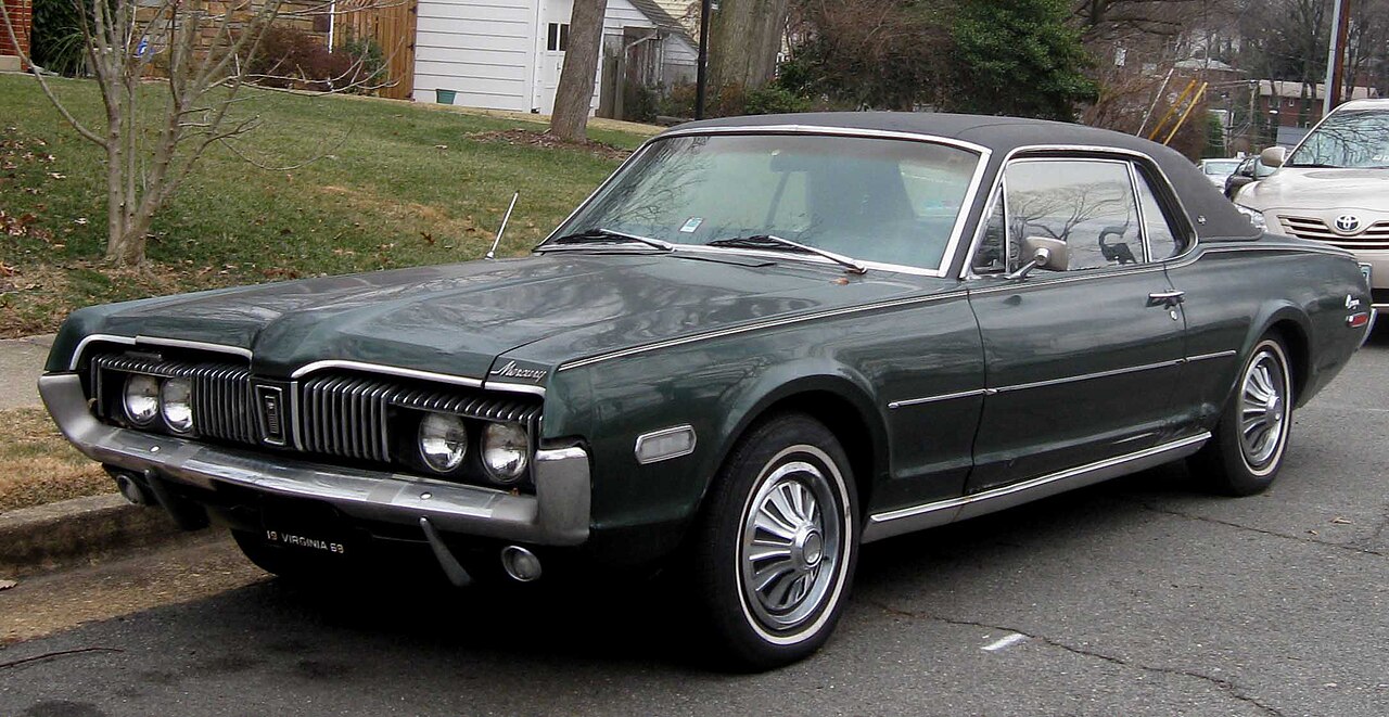 Dark green Mercury Cougar classic car parked on residential street. Vintage two-door coupe with distinctive chrome grille, elegant body lines, and period-correct wheel design, set against suburban backdrop