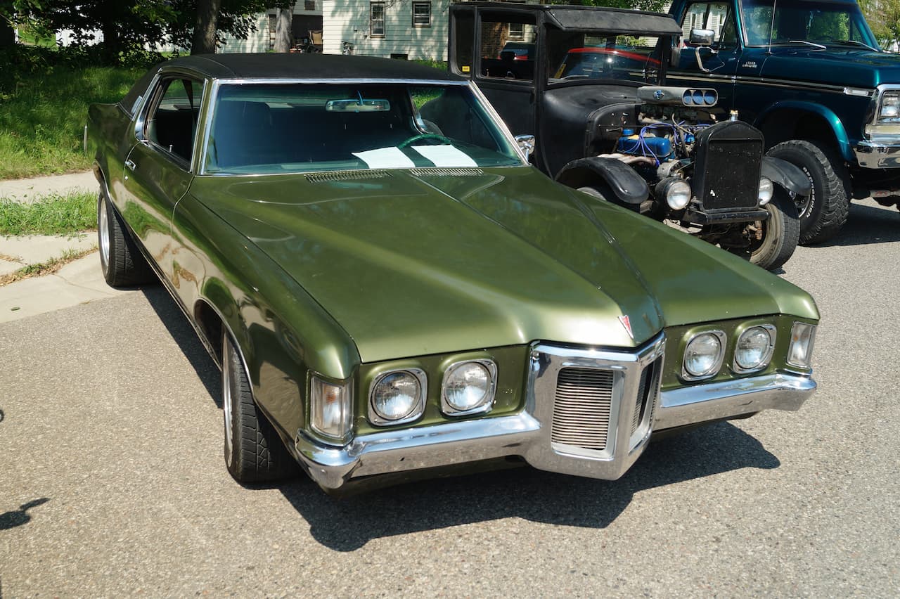 A dark green 1969 Pontiac Grand Prix with distinctive chrome front grille and quad headlights parked on a residential street. Behind it are a black vintage hot rod and part of a blue pickup truck
