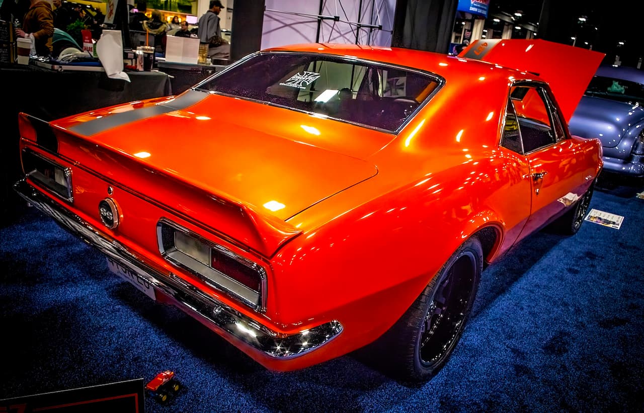 A bright orange 1969 Chevrolet Camaro with black racing stripes displayed at an auto show. The classic muscle car is photographed from the rear three-quarter angle, showcasing its glossy finish and chrome details against blue carpeting