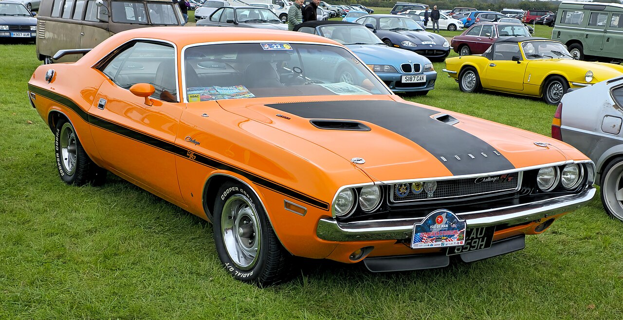 Bright orange Dodge Challenger with black hood stripe, parked on grass at a classic car show. Vintage muscle car with distinctive design, chrome details, and performance styling among other classic vehicles