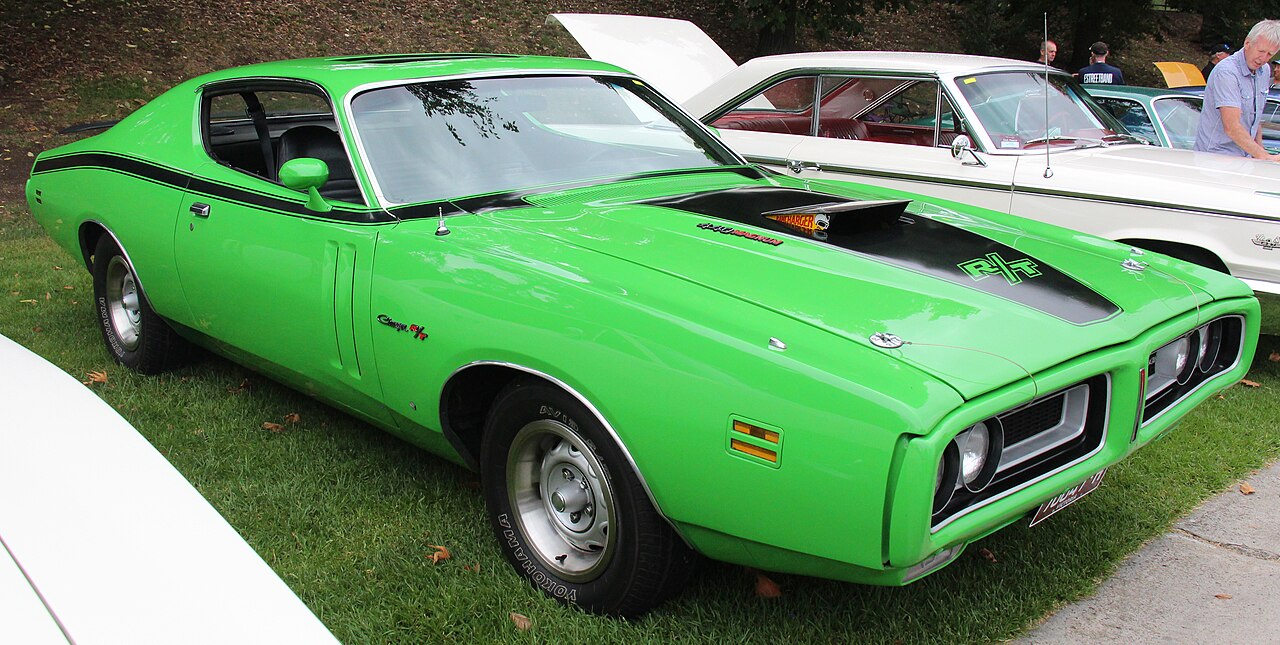 Bright green 1971 Dodge Charger R/T muscle car with black racing stripes and hood scoop displayed on grass at a car show. White classic cars visible in background with attendees browsing the exhibition