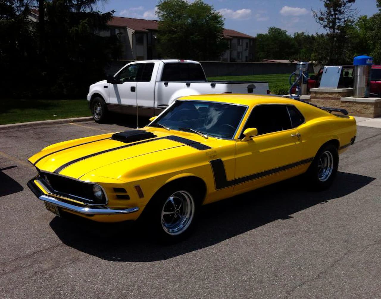 A bright yellow Ford Mustang Boss 302 with black racing stripes parked in a lot. The classic muscle car features distinctive side vents and chrome wheels. A white pickup truck is visible in the background with apartment buildings beyond