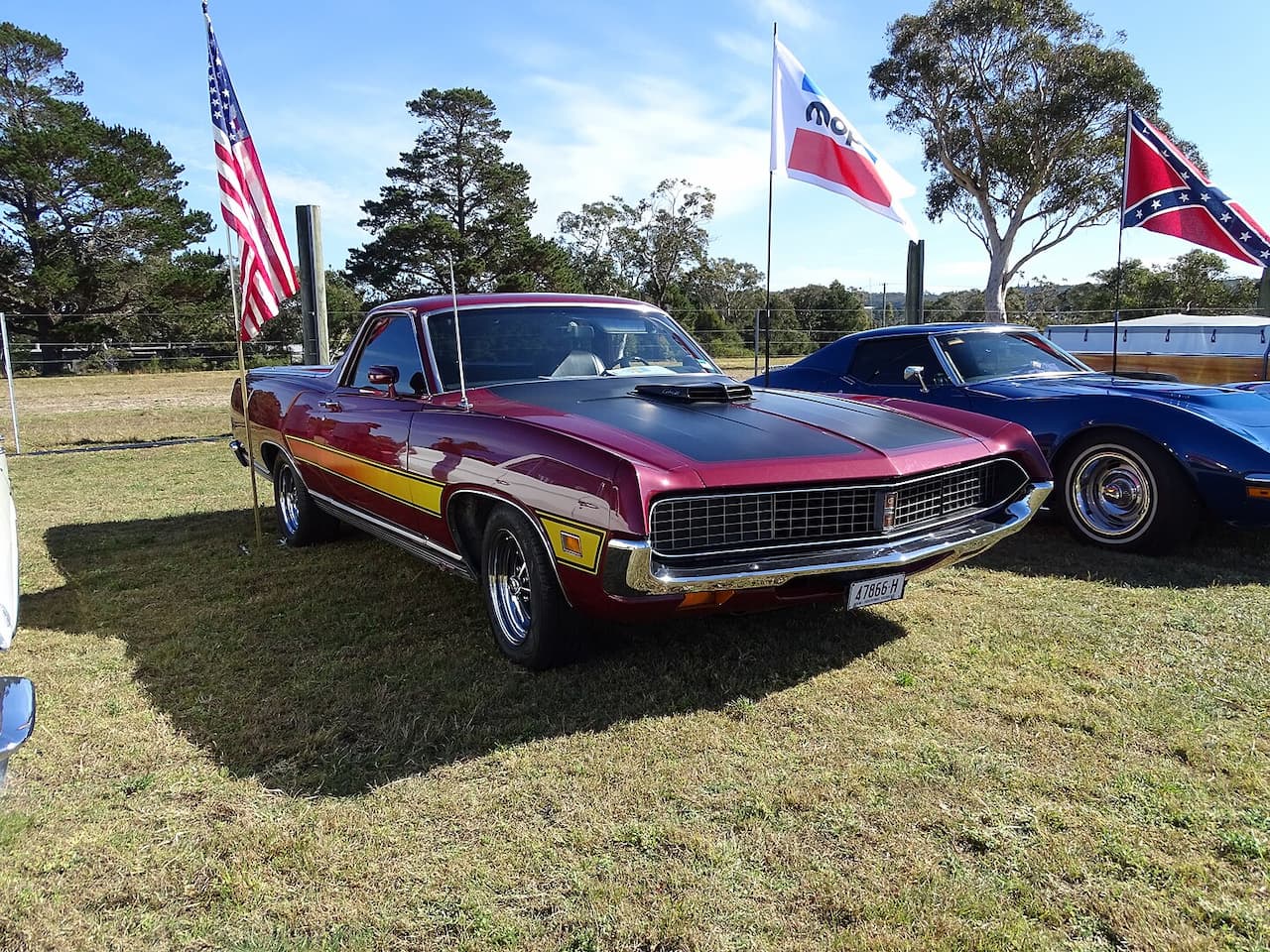 A burgundy 1971 Ford Ranchero GT with yellow side accents and a black hood scoop displayed at an outdoor car show. Several flags including American, Confederate, and state flags are mounted nearby. A blue classic car is partially visible to the right