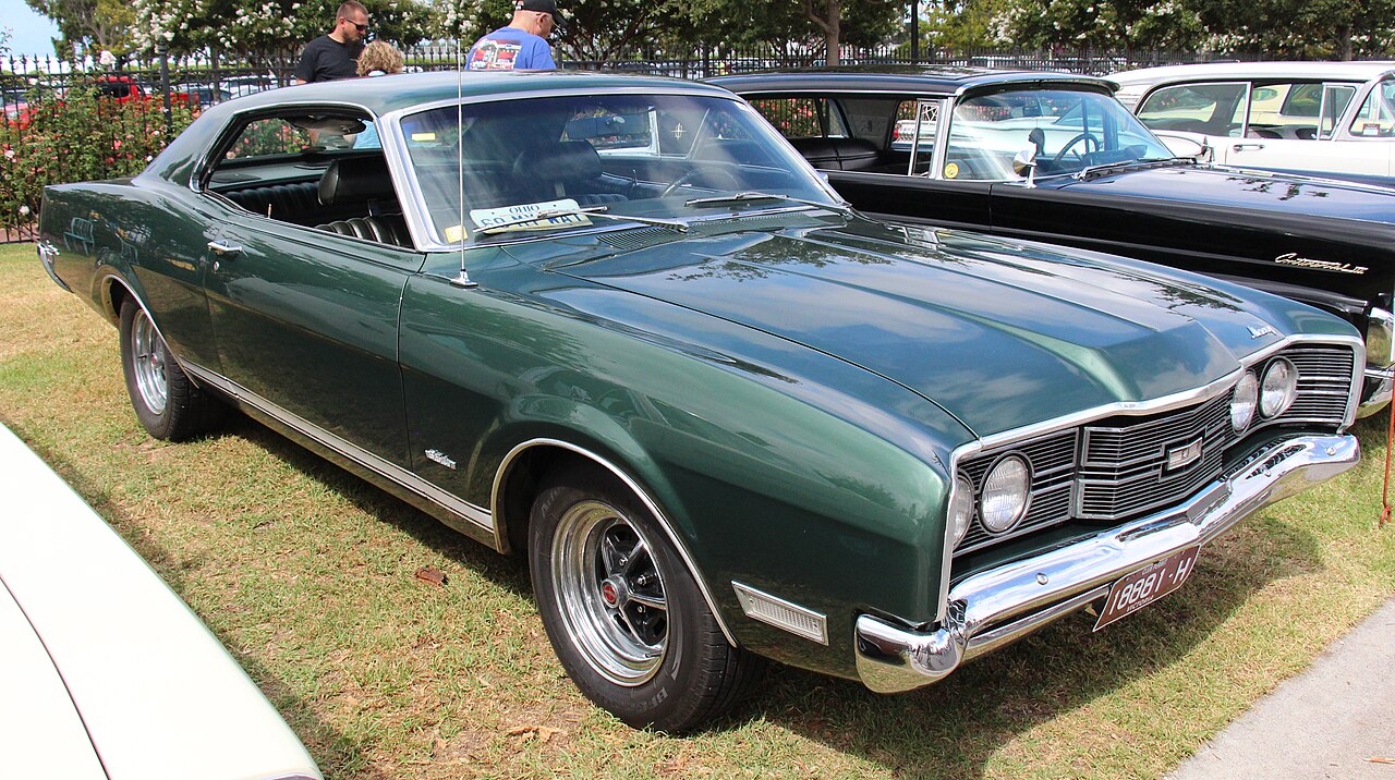 Green 1971 Mercury Cyclone Spoiler displayed on grass at car show. Features chrome trim, custom wheels, and vintage license plate. Other classic cars and spectators visible in background with flowering trees along perimeter fence