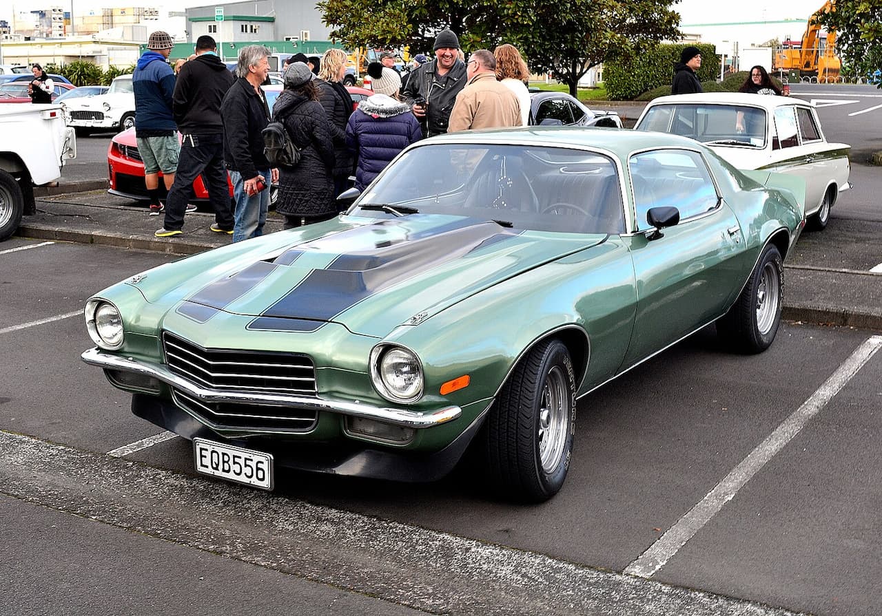 Metallic green vintage Chevrolet Camaro with dark racing stripes, parked at a car show. Crowd of people in winter clothing surrounds the classic muscle car in a parking lot setting