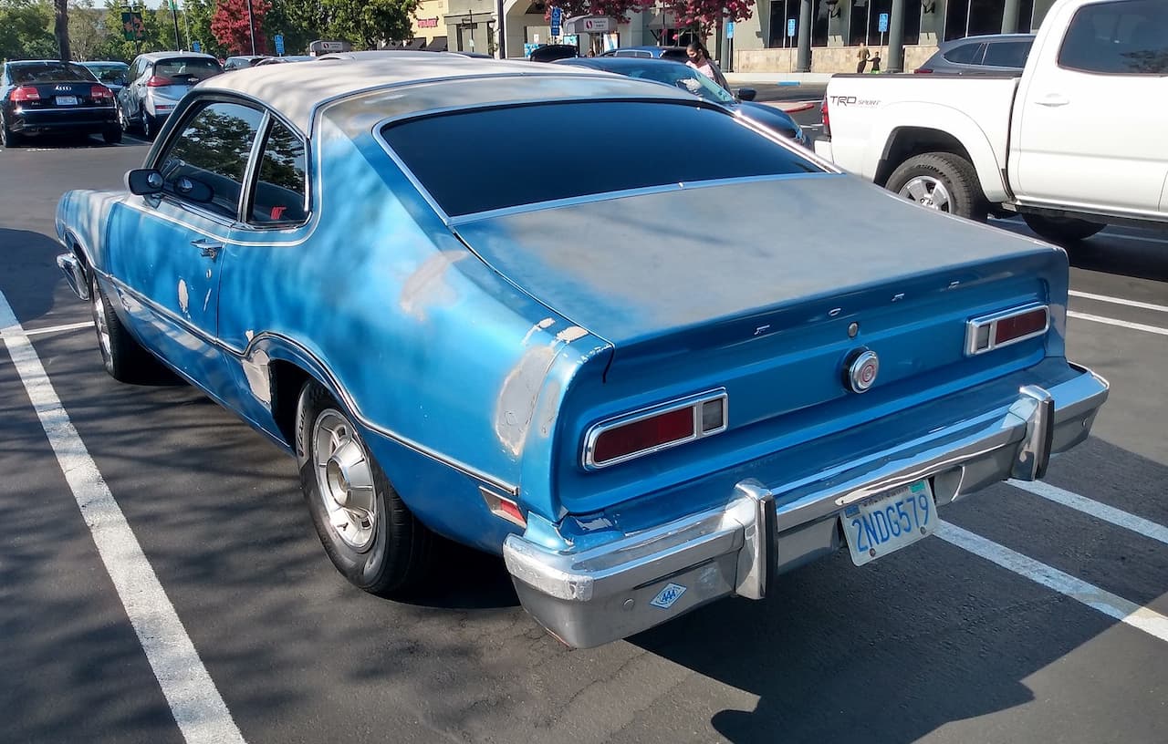 Vintage blue Ford Maverick parked in a shopping center lot. The classic sedan shows signs of wear with some body damage and mismatched panels. California license plate visible. Other modern vehicles parked nearby