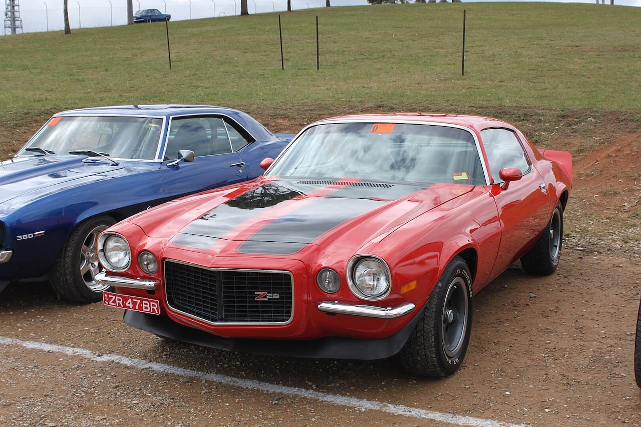 A red Chevrolet Camaro Z28 with black racing stripes and Australian license plate parked on dirt ground. A blue classic car, possibly a Camaro, is partially visible beside it. A grassy hill rises in the background