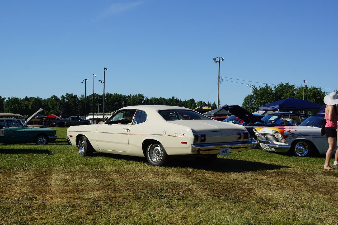 White 1974 Dodge Dart Hang10 displayed at an outdoor car show on grassy field. Other classic cars visible nearby under pop-up tents. Light poles and treeline in background under clear blue sky