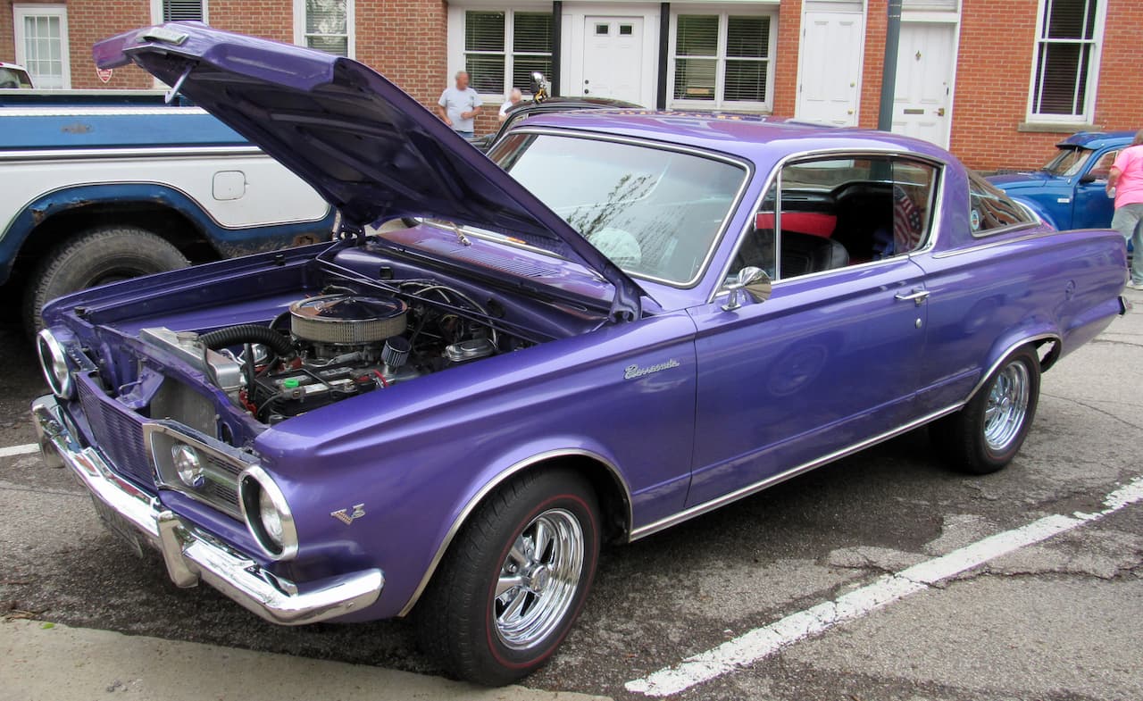A purple 67-69 Plymouth Barracuda classic car with its hood open, displaying the engine. The car is parked in front of a brick building, with chrome wheels and trim gleaming against its vibrant purple paint