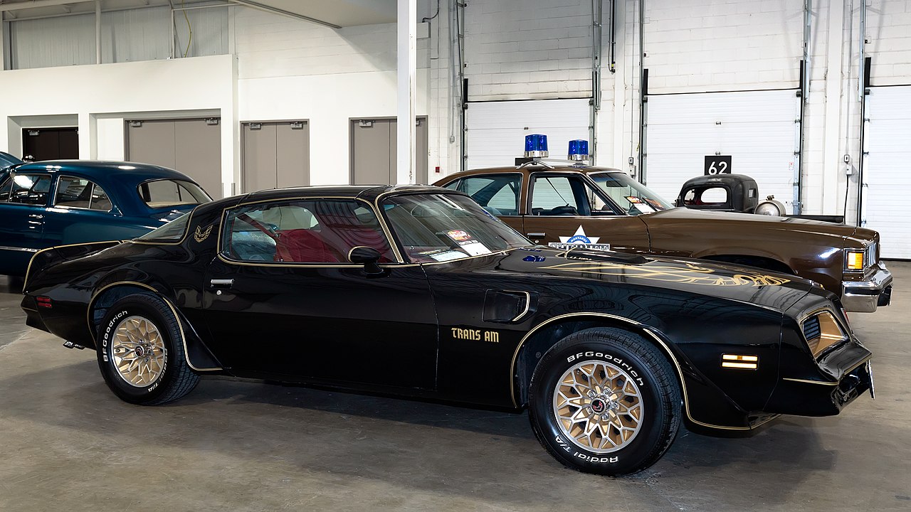Black Pontiac Can Am (1977) with gold accents and snowflake wheels displayed in warehouse garage. Red interior visible. Brown police cruiser with blue lights and other vintage vehicles parked nearby. Loading dock doors and room dividers in background