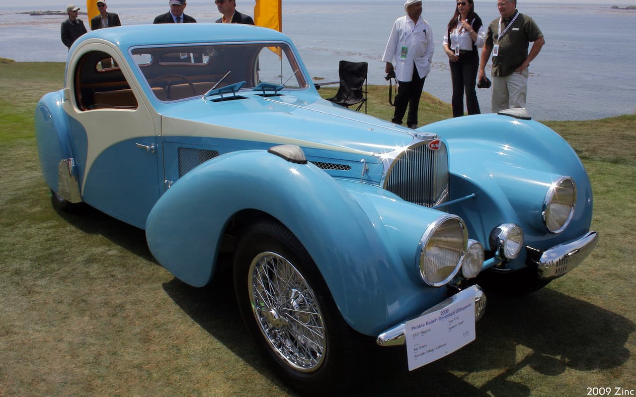 Vintage light blue Bugatti classic car with curved bodywork, displayed on grassy field at a car show. Gleaming chrome details, wire-spoke wheels, and multiple people in the background near a shoreline