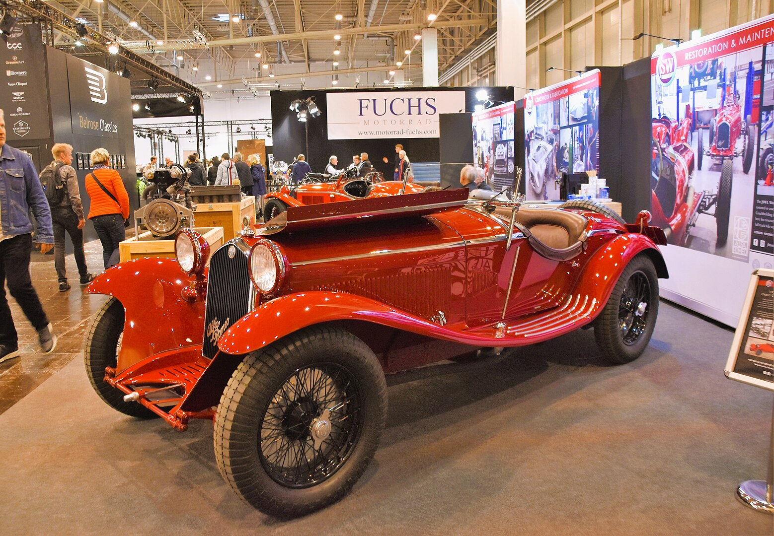 Vibrant red vintage classic car displayed at an indoor motor show. Sleek, open-top design with wire-spoke wheels, positioned in front of exhibition booths. People walking around, with "Fuchs Motorrad" sign visible in background