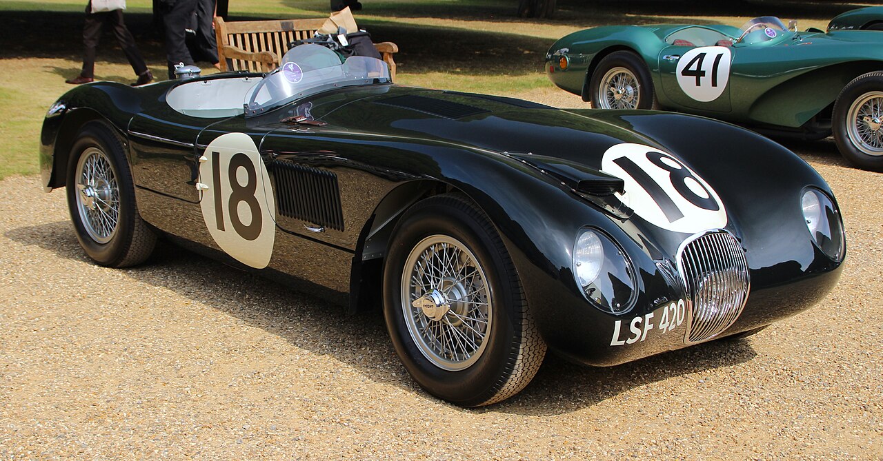 Classic black vintage racing Jaguar sports car with racing number 18, featuring wire-spoke wheels, positioned on gravel or sand at an event. Sleek curved bodywork, prominent grille, and another green racing car partially visible in background