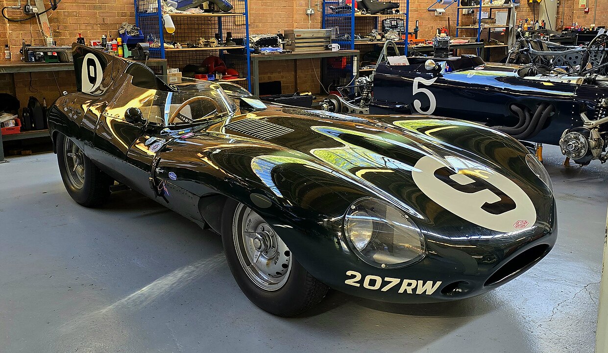 Sleek black vintage racing car with number 9, parked in a workshop. Reflective bodywork shows detailed curves, with license plate 207RW. Brick walls, blue mesh shelving, and various mechanical equipment visible in background