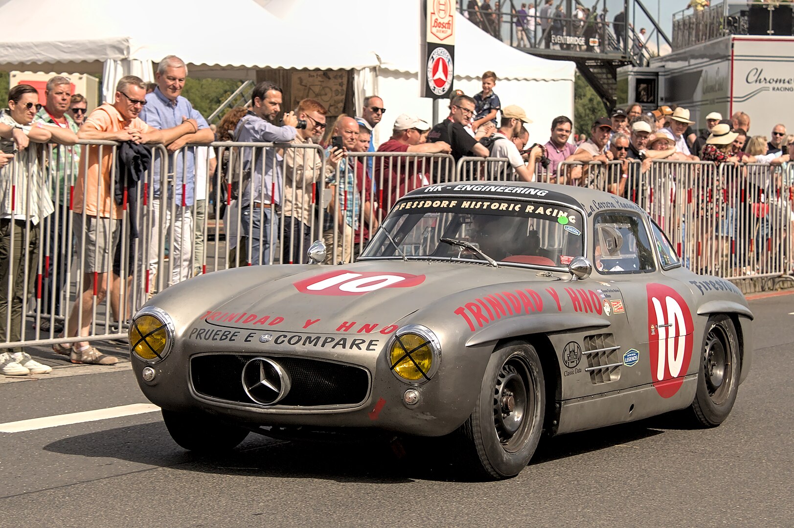A vintage silver Mercedes-Benz 300SL race car with red decals and the number "10" is seen driving past a crowd at a classic car event. The car features yellow-tinted headlights, a large front grille, and historic racing stickers on its body