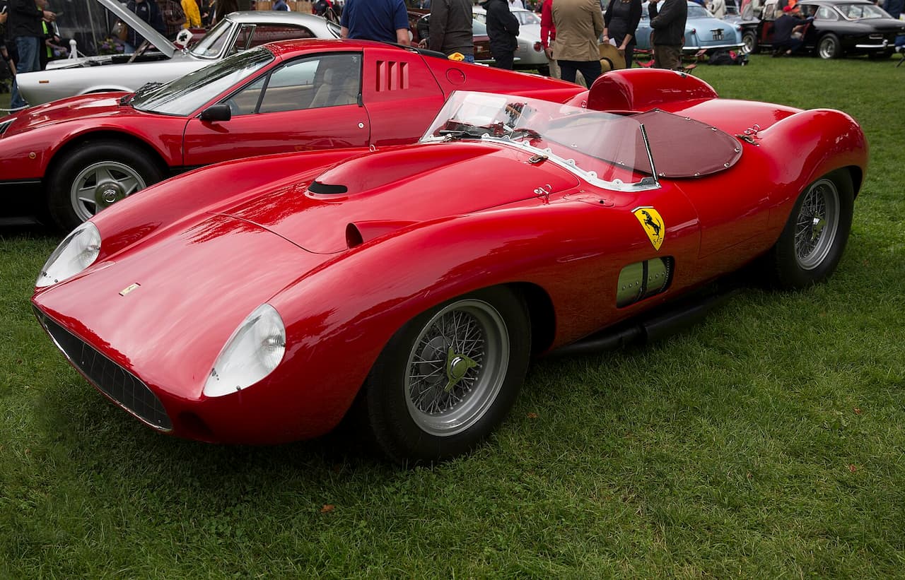 A classic red Ferrari race car with an open cockpit is displayed on a grassy field. The sleek body features wire-spoke wheels, a transparent windscreen, and Ferrari’s prancing horse emblem. Other vintage cars and spectators are visible in the background
