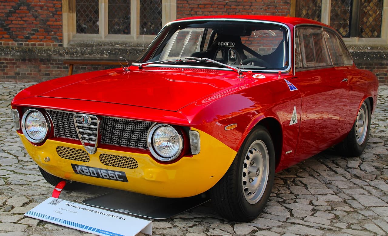 A red and yellow 1965 Alfa Romeo Giulia Sprint GTA is displayed on a cobblestone surface. The classic race car features round headlights, a distinctive Alfa grille, Sparco racing seats, and Quadrifoglio badges on the fenders