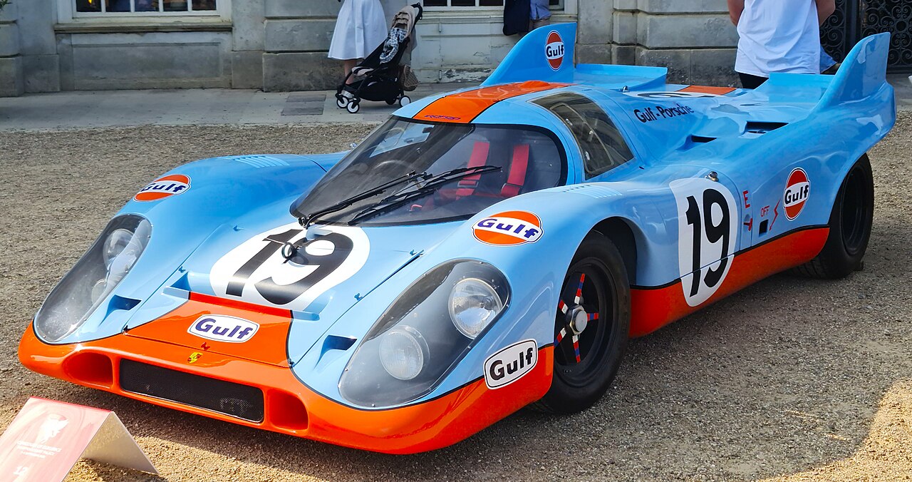 A Porsche 917K race car in iconic Gulf Oil livery, featuring a light blue and orange color scheme with the number "19," is displayed on gravel. It has a sleek aerodynamic design, large headlights, and a curved windshield, with spectators nearby