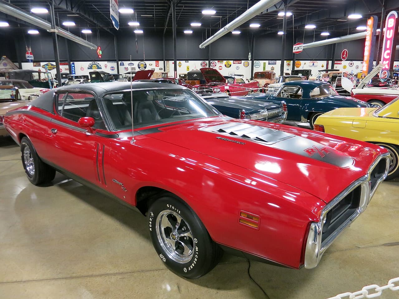 A cherry red 1971 Dodge Charger R/T with black roof and chrome wheels displayed at an indoor classic car museum or showroom. Multiple vintage vehicles are visible in the background beneath industrial ceiling lights