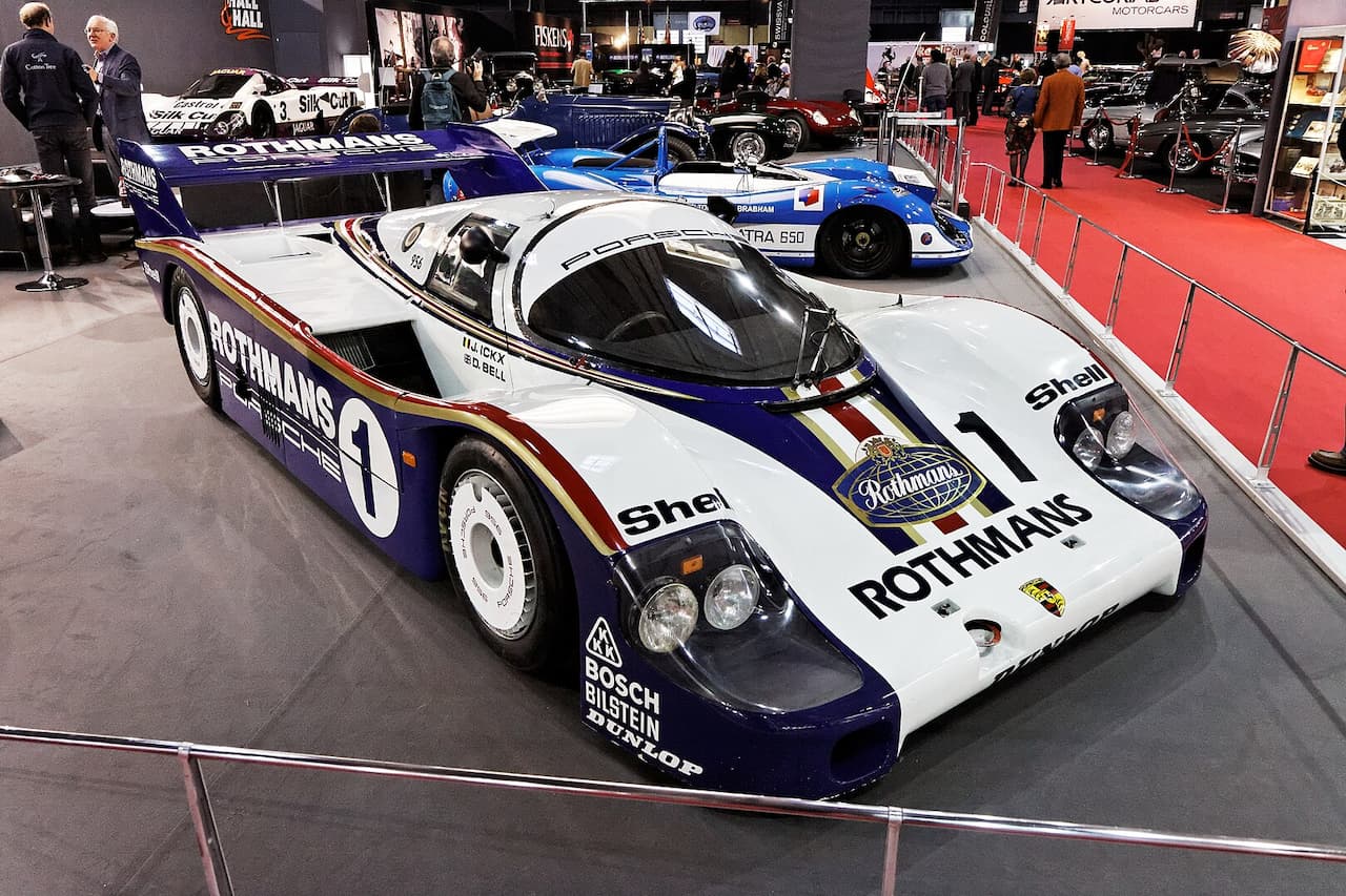 A Porsche 956 race car in Rothmans livery is displayed at an auto show. The sleek white, blue, and red body features sponsor logos, a large rear wing, and a bubble canopy. Other vintage race cars and spectators surround the exhibition area