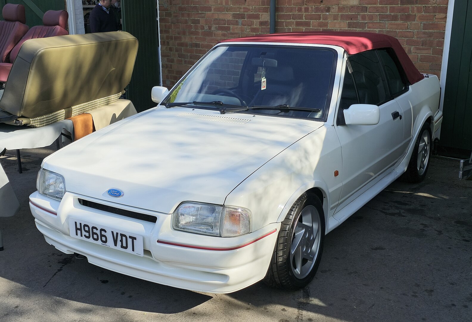 White Ford convertible with red soft top, parked in front of a brick wall. Silver wheels, clear headlights, British license plate. Vintage car with classic 1980s-90s design, side view showcasing automotive styling