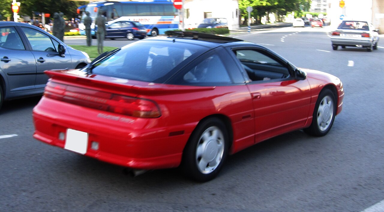 Red sports car with black roof, likely a Mitsubishi 3000GT, driving on a city street. Multiple vehicles and urban surroundings visible. Blurred motion suggests active traffic scene
