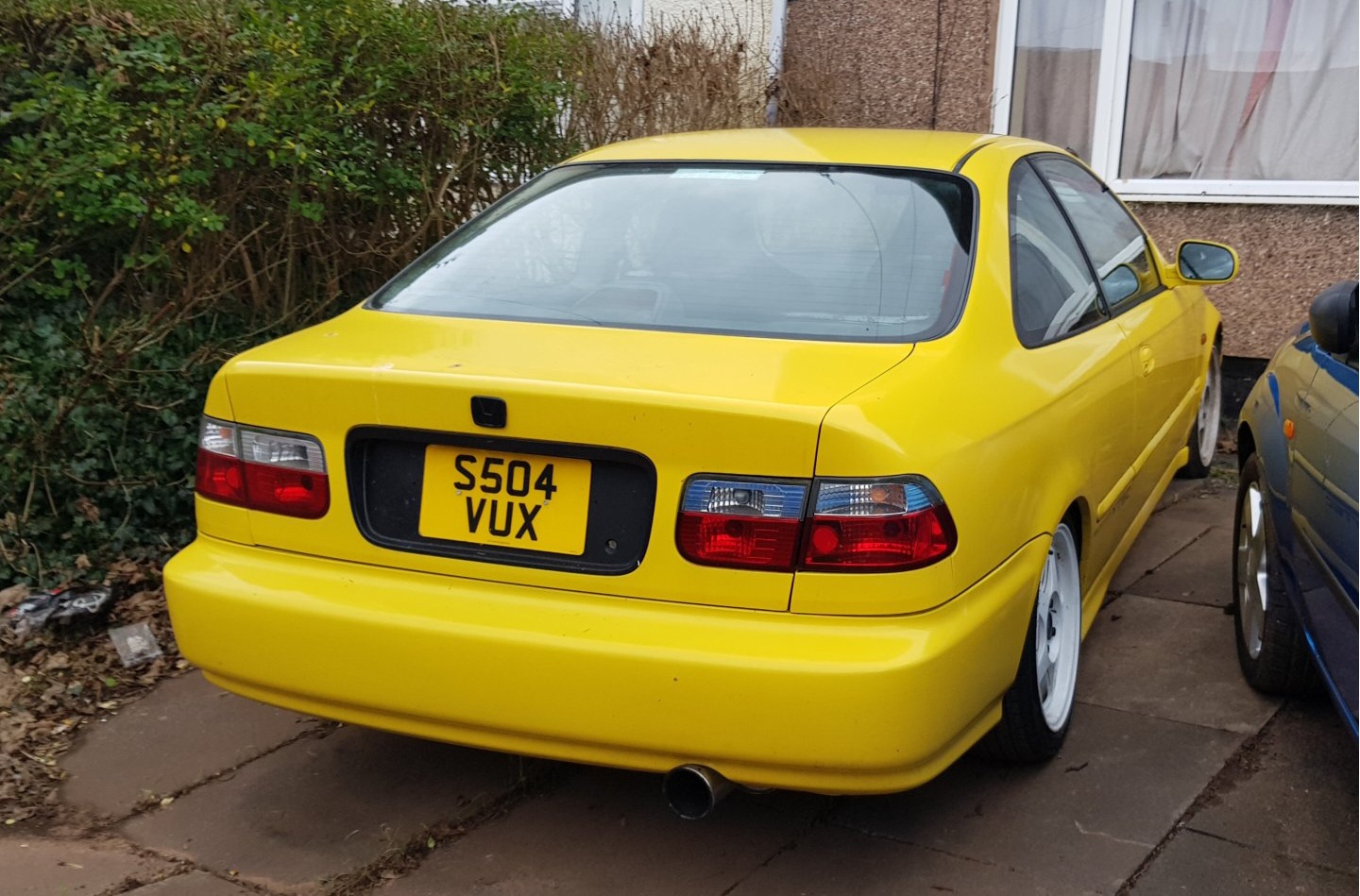 Bright yellow Honda Civic coupe parked on a concrete surface. Red taillights, white wheels, and a UK license plate (S504 VUX) are visible. Surrounding area includes a hedge and building wall