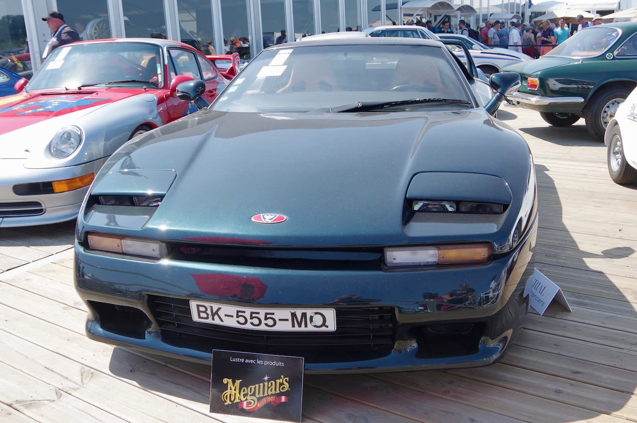 Dark blue sports car at automotive show. Vintage model with pop-up headlights, displayed on wooden platform with Meguiar's car care product sign. Multiple vehicles and crowd visible in background