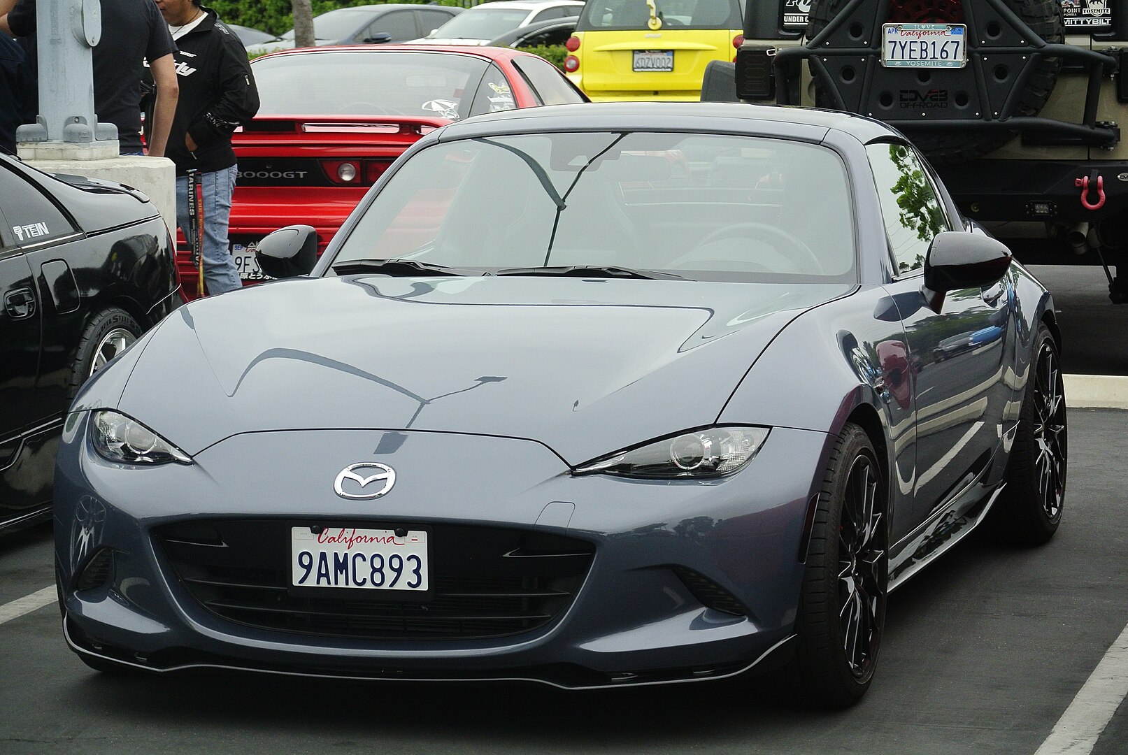 A gray Mazda MX-5 Miata with a California license plate is parked at a car meet. The sports car has sleek headlights, black alloy wheels, and a low stance. Other vehicles, including a red Mitsubishi 3000GT and a black off-road SUV, are visible in the background