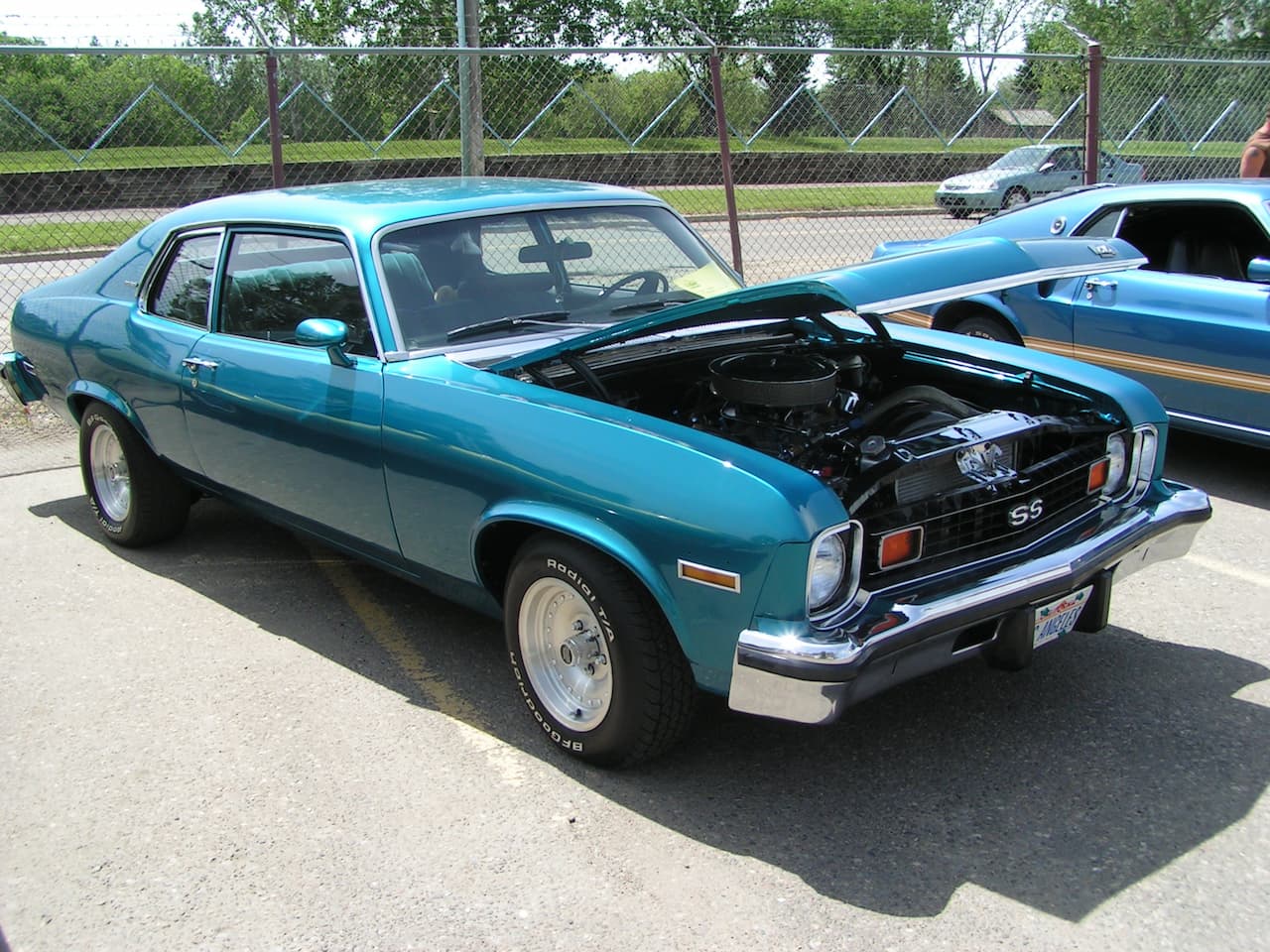A teal blue Chevrolet Nova SS with its hood open, displaying the engine. The classic muscle car is parked on pavement at what appears to be a car show or meet, with a chain-link fence and trees in the background