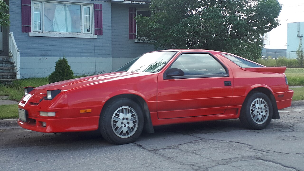 A red Dodge Daytona sports coupe parked on a cracked asphalt street. The two-door car features white alloy wheels and a rear spoiler. Background shows a blue house with burgundy shutters and green trees