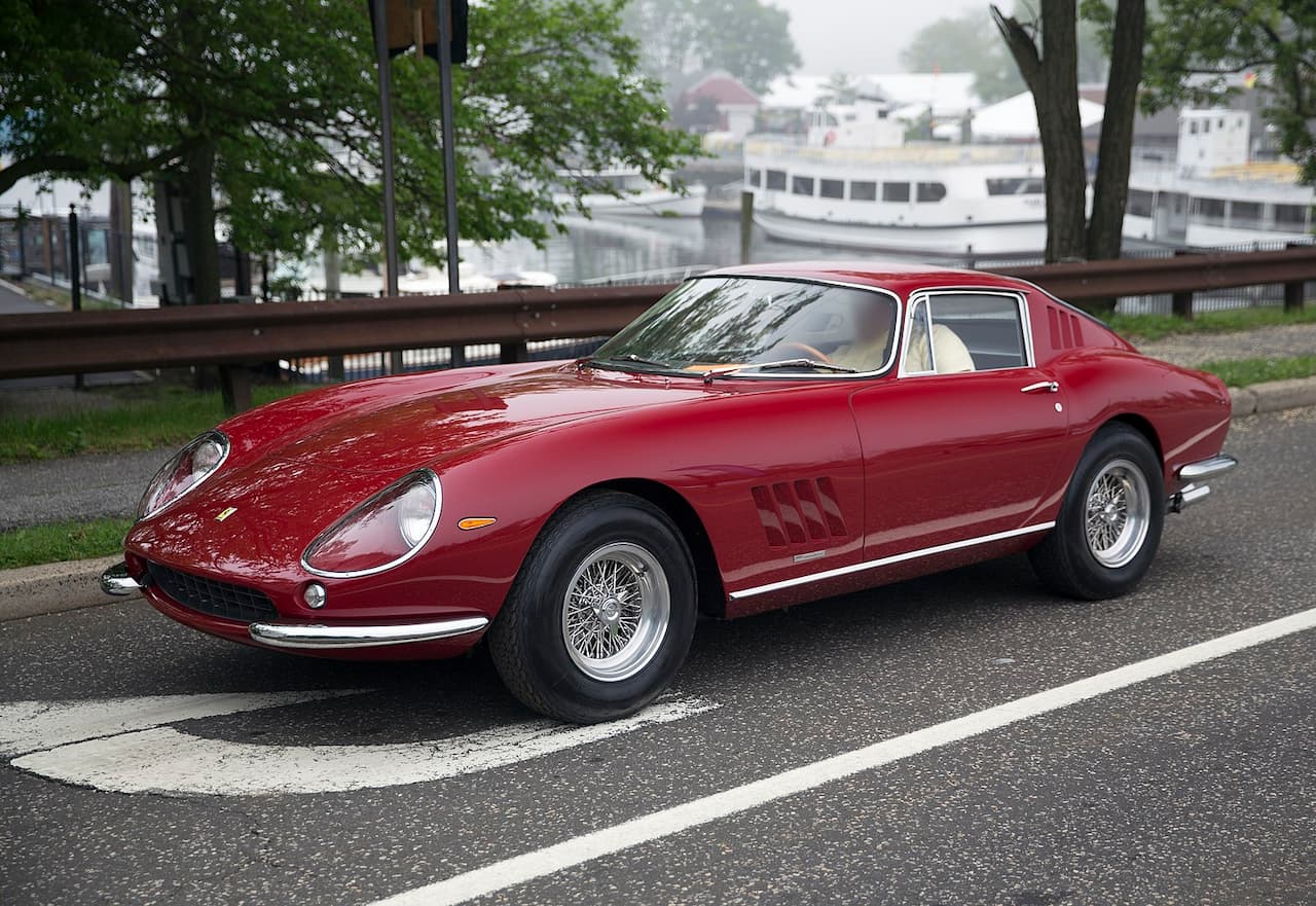 A classic red Ferrari 275 GTB is parked on a road with white markings. The car features a sleek, curvy design, round headlights, chrome bumpers, wire-spoke wheels, and cream-colored interior seats. A waterfront with boats and trees is visible in the background