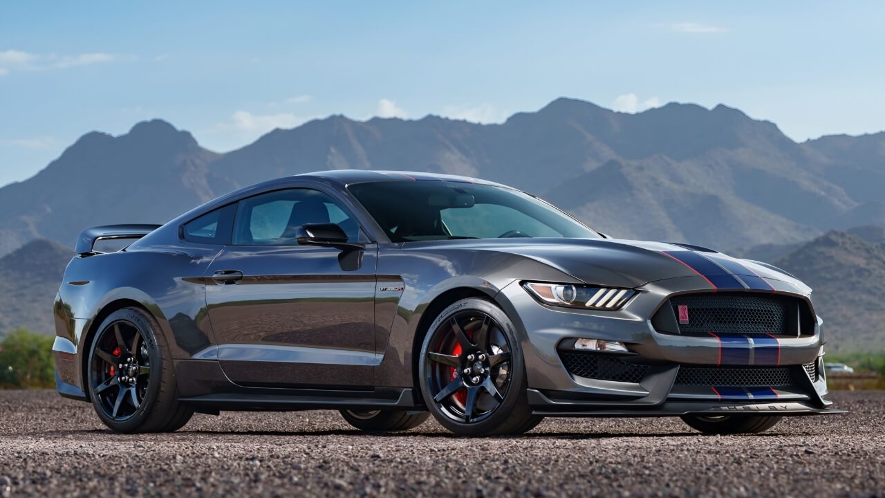 Dark gray Ford Mustang Shelby GT350R with racing stripes and aerodynamic spoiler parked against mountain landscape backdrop