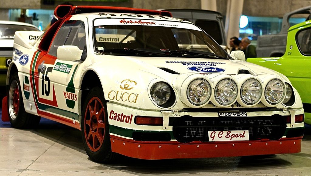 White and red Ford racing car with multiple sponsor logos, featuring four additional headlights, and displayed in an indoor exhibition setting with a green car partially visible