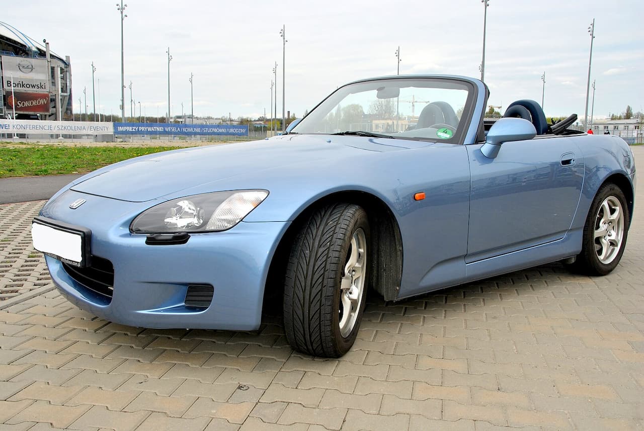 Light blue Honda S2000 convertible sports car parked on brick-paved surface. Soft top down, showing sleek design with modern headlights. Background includes industrial-looking structures and signage