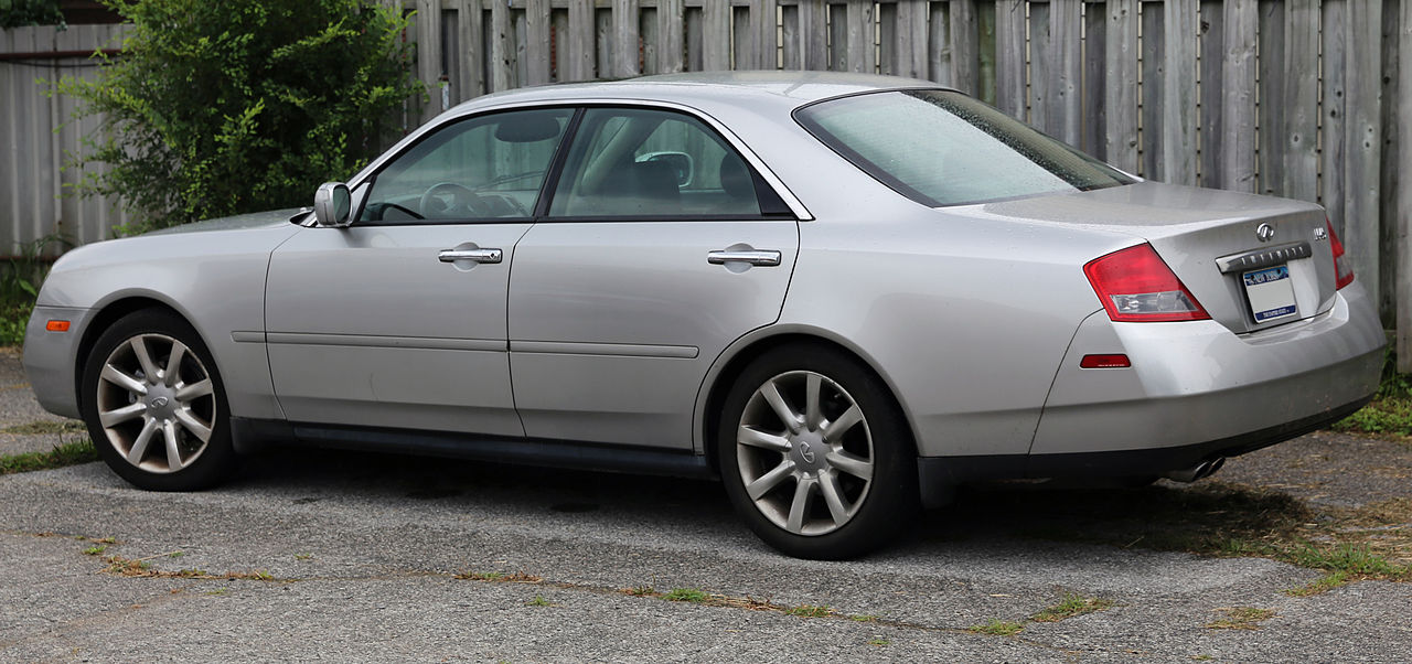 Silver Infiniti M-series sedan parked on a cracked asphalt surface, situated next to a weathered wooden fence with green foliage in the background. The car is shown from a three-quarter rear view