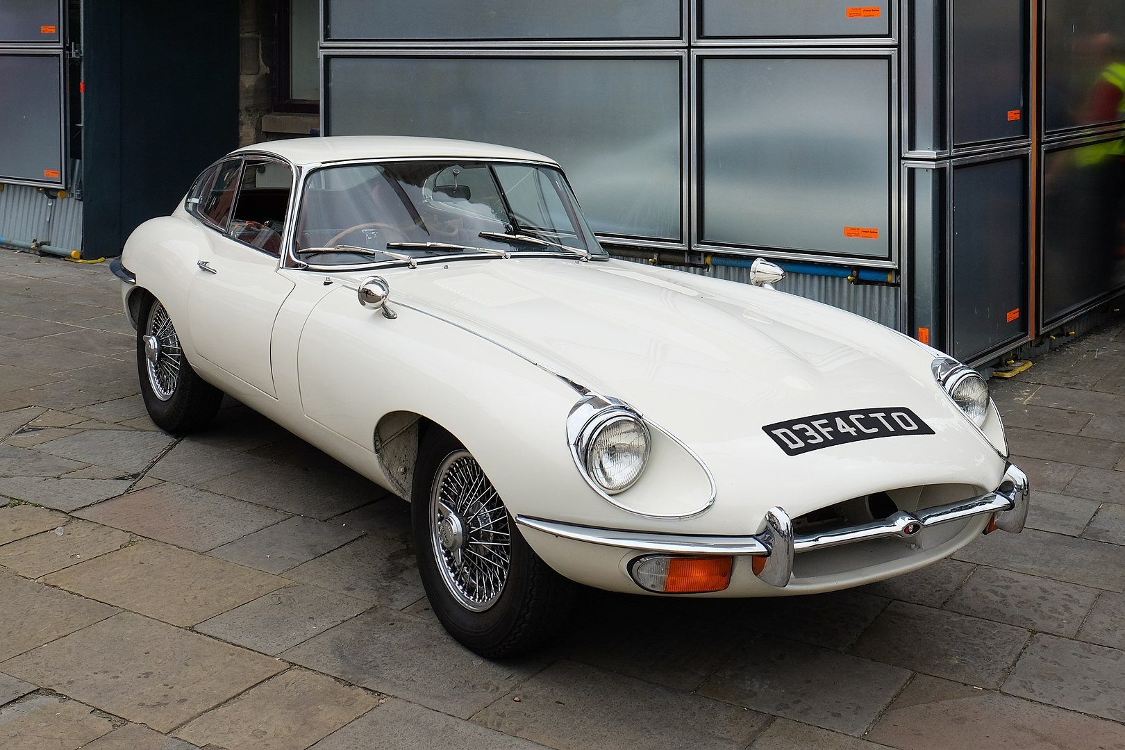 A classic white Jaguar E-Type Series 2 coupé is parked on a stone pavement. The car features smooth curves, chrome bumpers, round headlights, and wire-spoke wheels