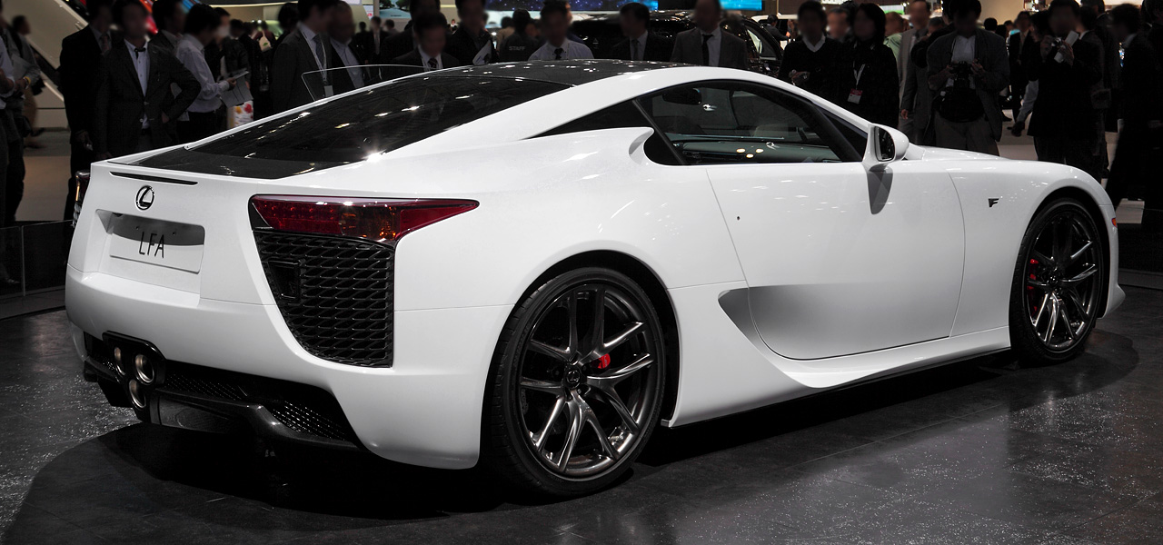 A white Lexus LFA sports car is displayed at an auto show, surrounded by a crowd. The rear showcases its triple exhaust, carbon fiber accents, and aggressive taillights. The black roof and large alloy wheels enhance its aerodynamic, high-performance design