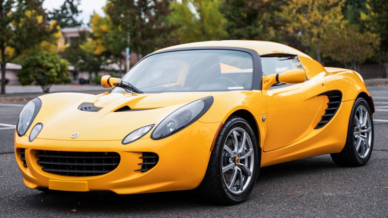 Bright yellow Lotus Elise parked on asphalt with trees and buildings visible in background