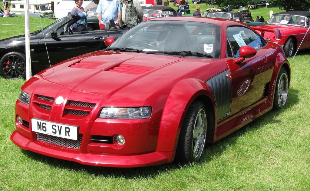 Vibrant red sports car (MG XPower SV) displayed at outdoor car show. Aggressive design with mesh grille, muscular body lines, parked on green grass with other vehicles and spectators in background.
