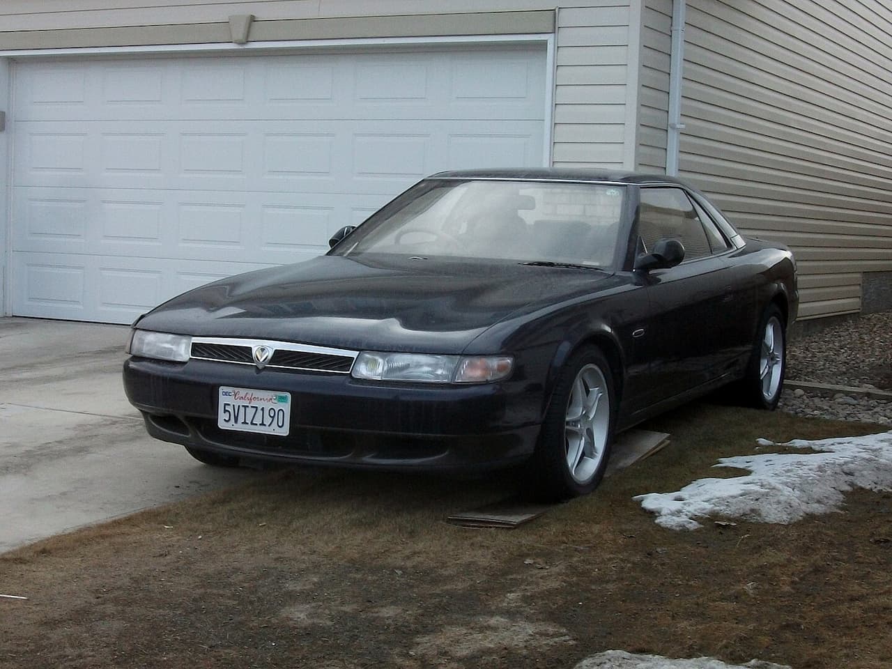 Black Mazda 626 coupe parked in front of a white garage door, with California license plate. Sits on clean alloy wheels with snow and dirt surrounding the parking area