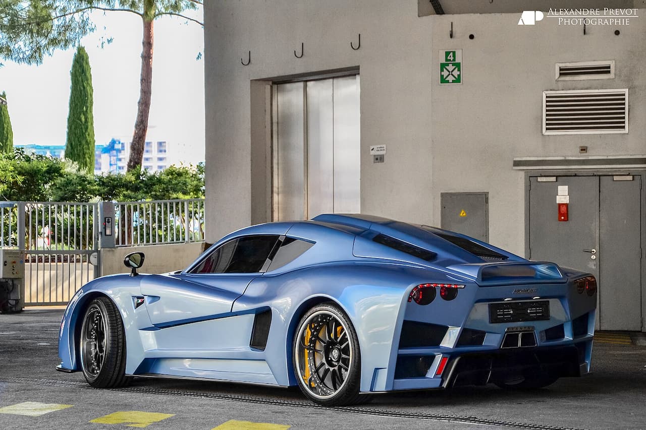 Blue-silver supercar parked in urban parking area. Sleek, angular design with distinctive rear air intakes, yellow brake calipers, positioned near building with safety signage and cypress trees in background