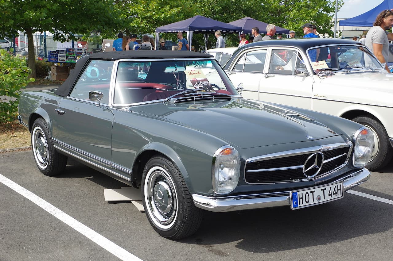 A classic Mercedes-Benz 230 SL Pagoda in grey with a black soft top is parked at an outdoor event. The vintage roadster features chrome bumpers, a Mercedes grille, round headlights, with people and other classic cars in the background
