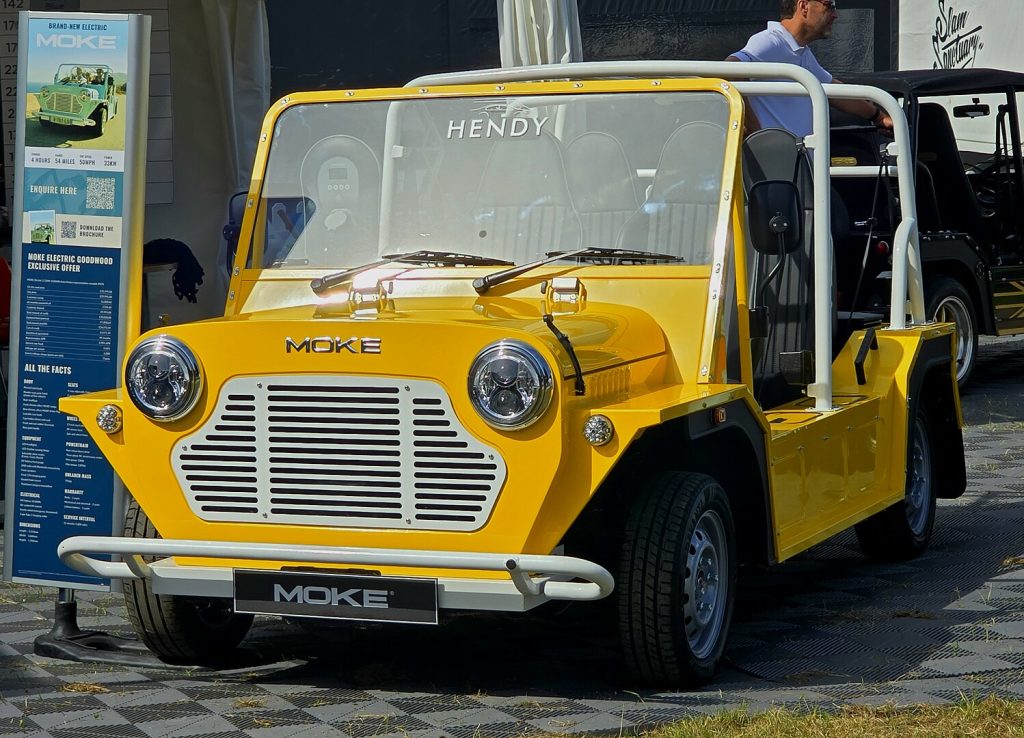 Yellow Mini Moke electric vehicle with "Hendy" branding, displayed at showroom. Modern compact convertible with distinctive round headlights, white roll cage