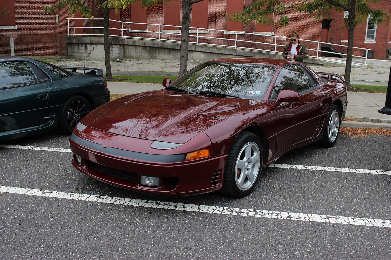 A maroon Mitsubishi 3000GT VR-4 sports car is parked in an outdoor lot with white-striped pavement. It features pop-up headlights, silver five-spoke wheels, and a sleek body. A dark green car, a brick building, and a woman using her phone are in the background