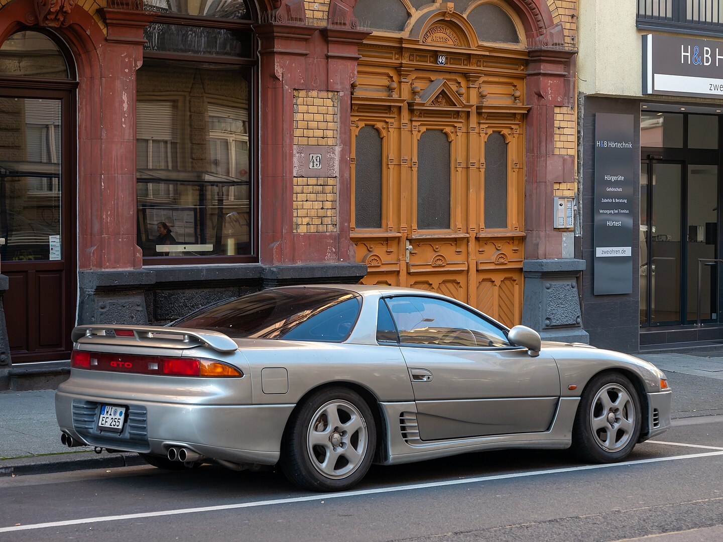 A silver Mitsubishi GTO is parked on a city street in front of an ornate wooden door and a brick building. The car features a rear spoiler, quad exhaust tips, and a sleek, aerodynamic design, reflecting the surrounding architecture in its glossy paint